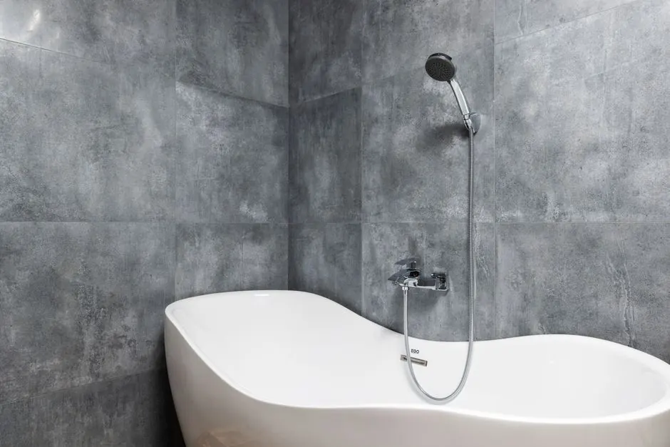 Interior of light modern bathroom with white bath under shower on gray tile wall